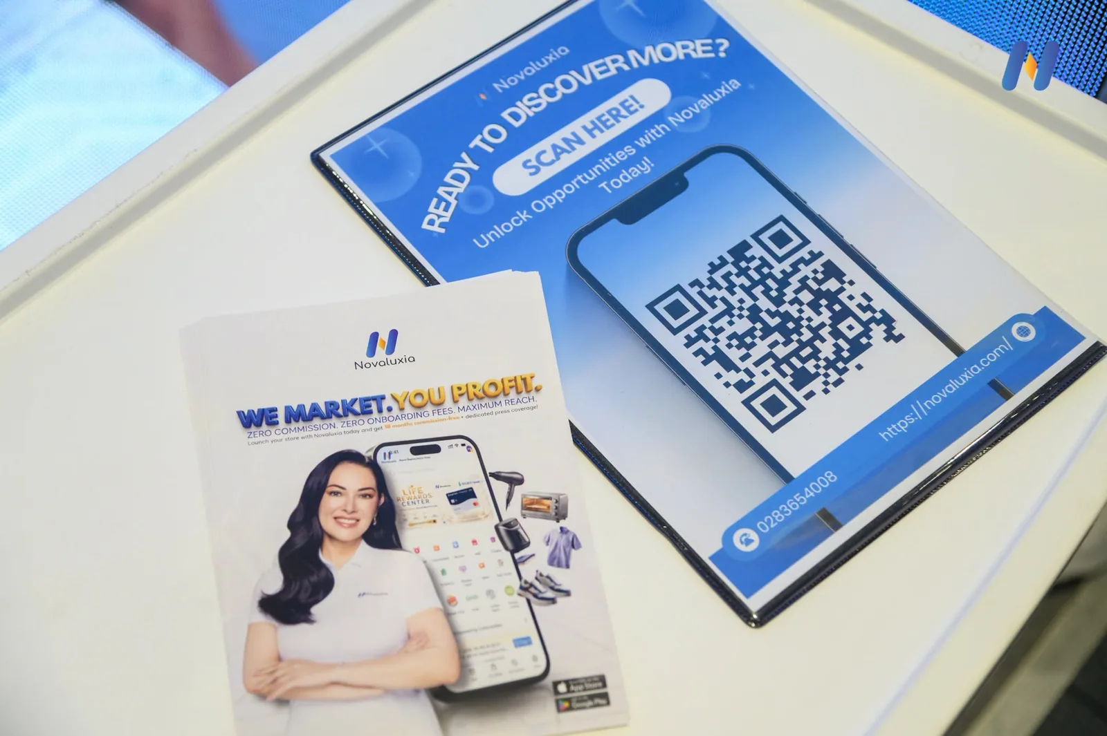 Novaluxia app table card printed with the line READY TO DISCOVER MORE? SCAN HERE! above a QR code at the booth