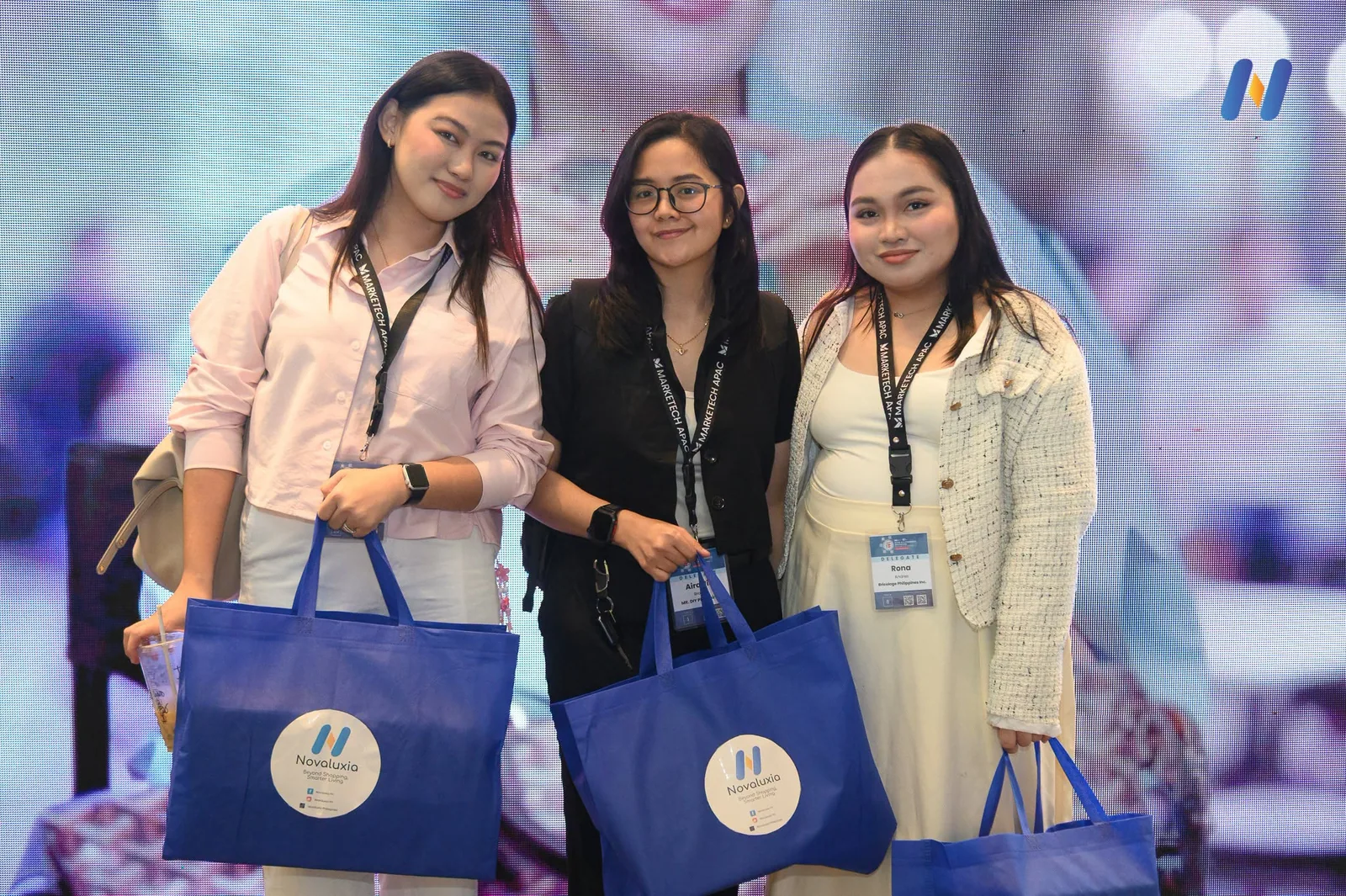 Three people holding up blue Novaluxia tote bags at the booth
